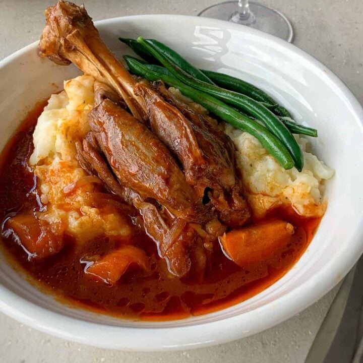 A bowl of braised lamb shank with sauce, served over mashed potatoes and garnished with green beans.