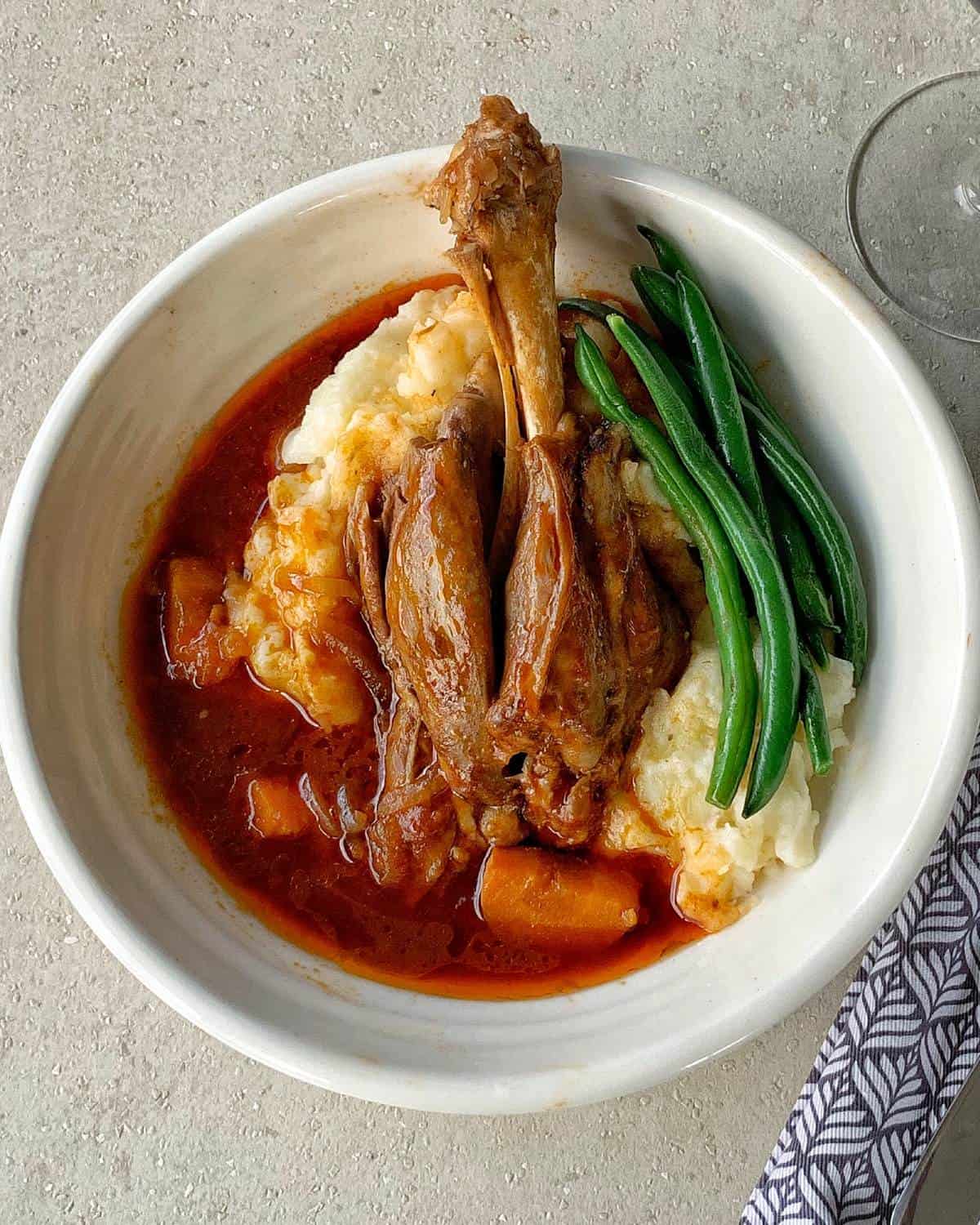 A bowl with braised lamb shank in sauce, served over mashed potatoes with cooked green beans on the side.