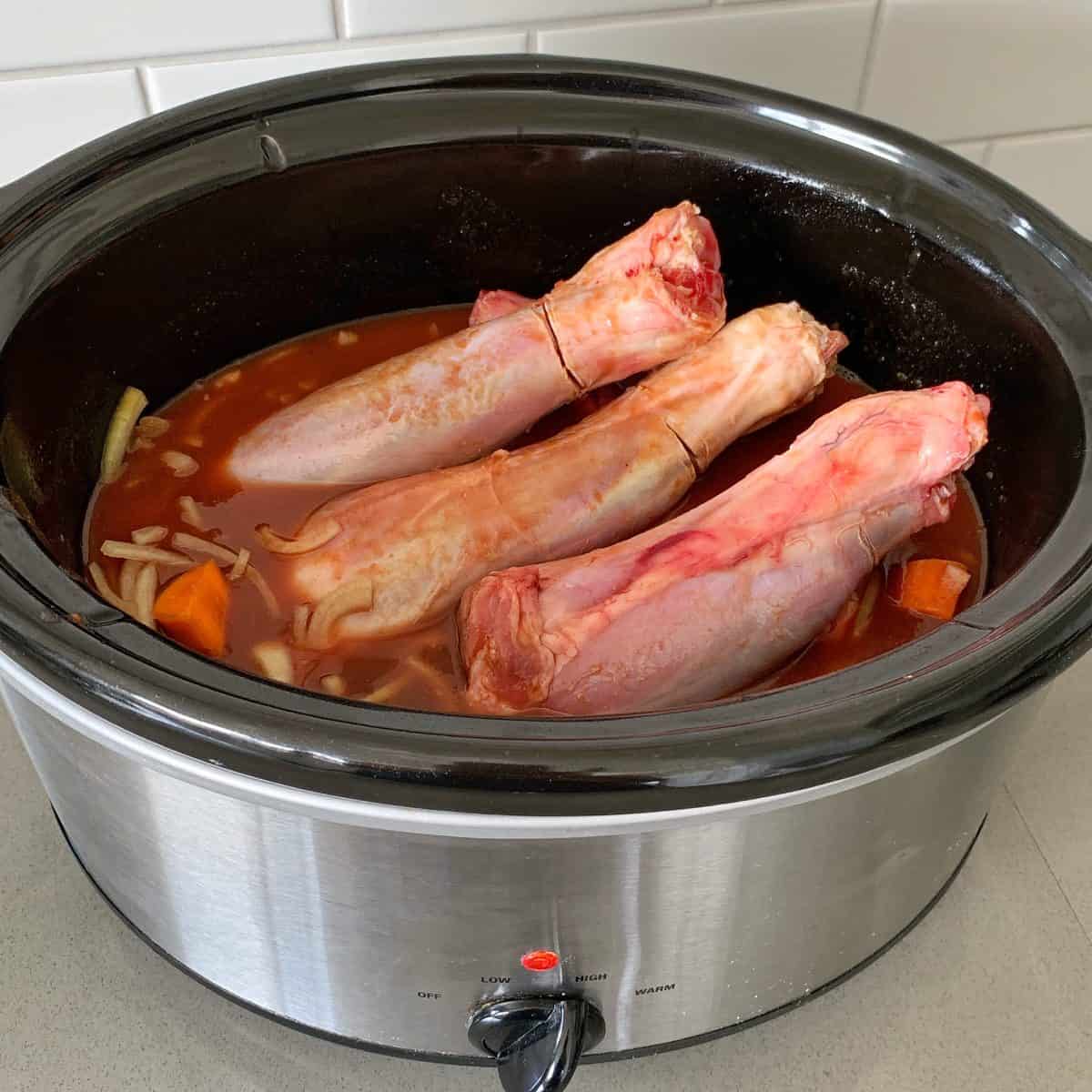 Three raw beef shanks in a slow cooker with broth, chopped vegetables, and seasonings, set on a kitchen counter with white tile backsplash.