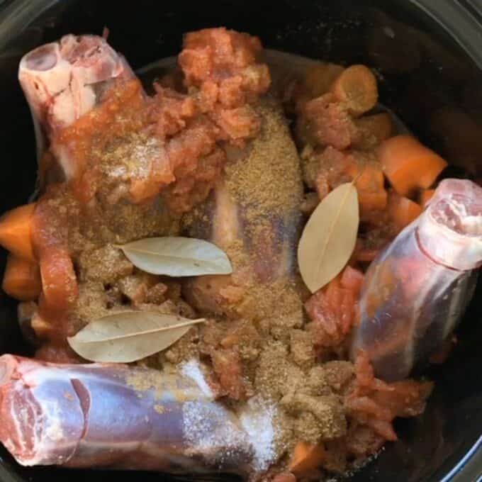 Three raw lamb shanks, chopped carrots, diced tomatoes, spices, and bay leaves in a slow cooker before cooking.