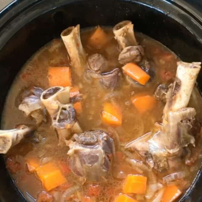 A slow cooker filled with a stew of lamb shanks, diced carrots, onions, and a brown broth.