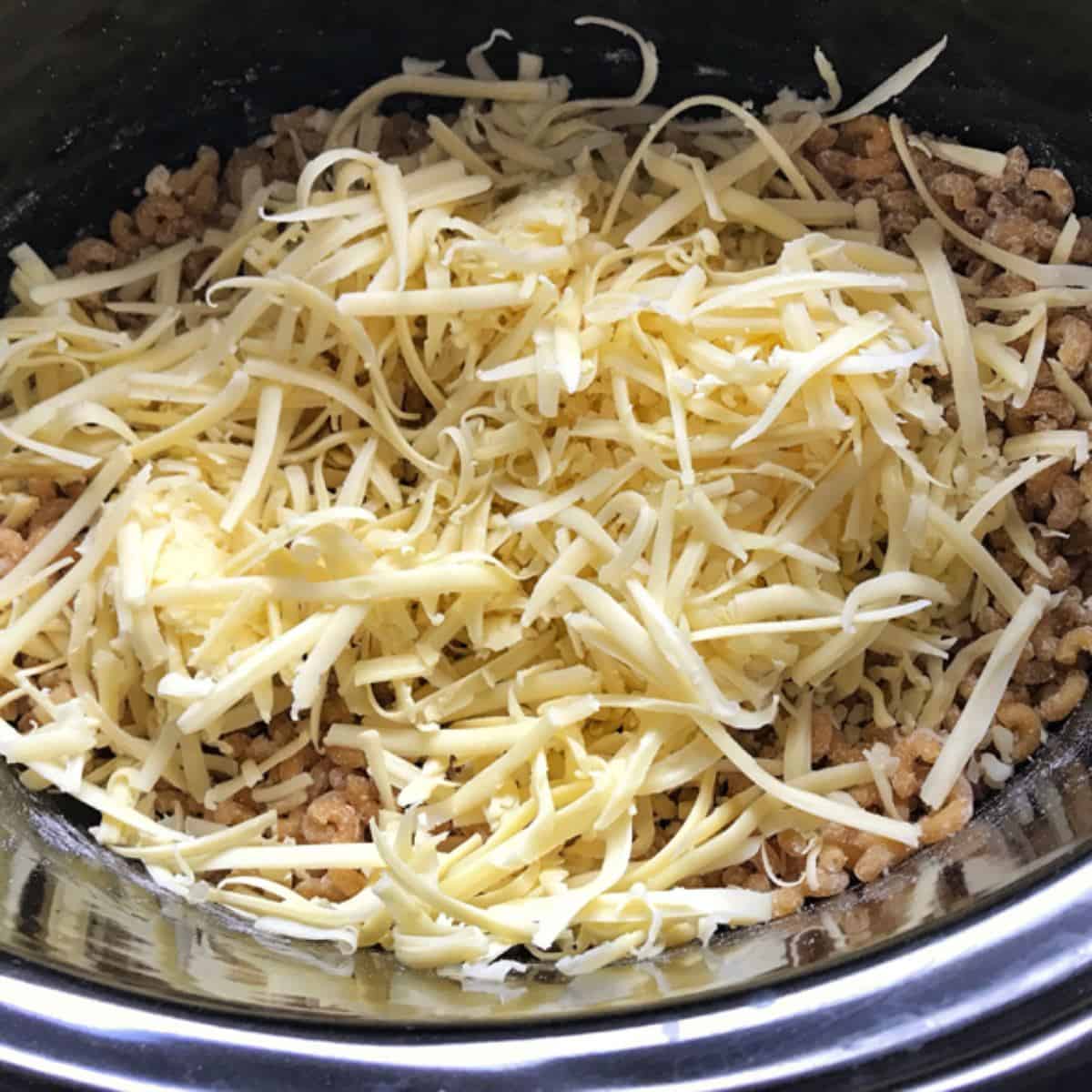 Slow Cooker Mac and Cheese Shredded cheese on top of cooked ground meat in a black pot or slow cooker.