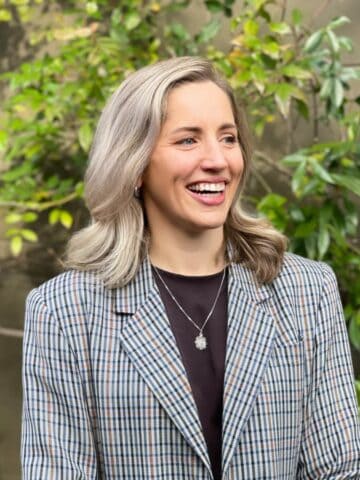 A woman with shoulder-length blonde hair, wearing a plaid blazer and necklace, stands outdoors in front of green foliage, smiling and looking to the side.
