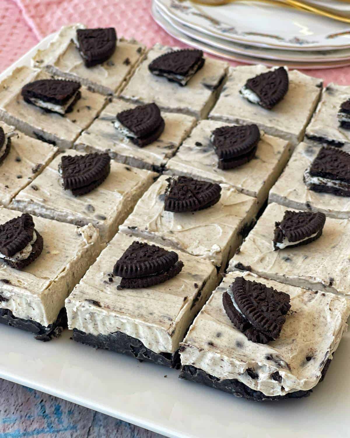 A plate of sixteen Oreo cheesecake bars, each topped with a piece of Oreo cookie, displayed on a white rectangular dish.