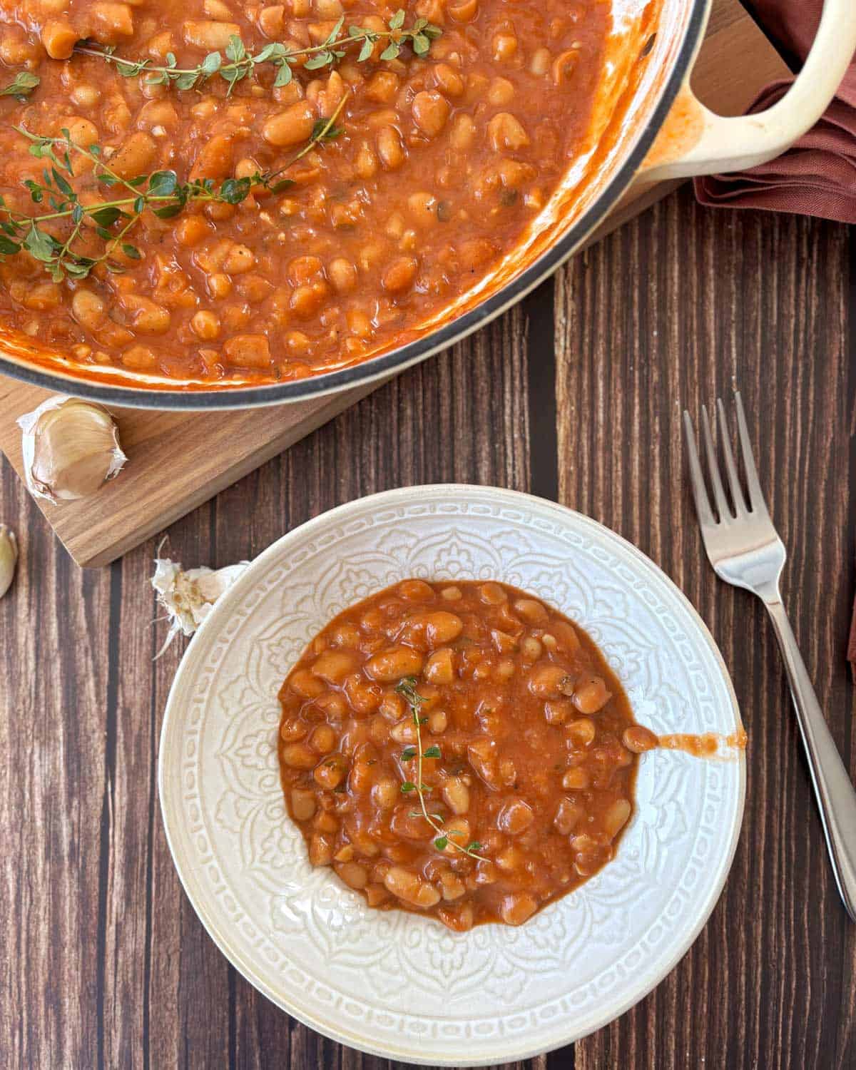 Homemade Baked Beans A bowl of baked beans in tomato sauce garnished with a sprig of thyme, next to a pot of beans on a wooden table with a fork and garlic cloves nearby.
