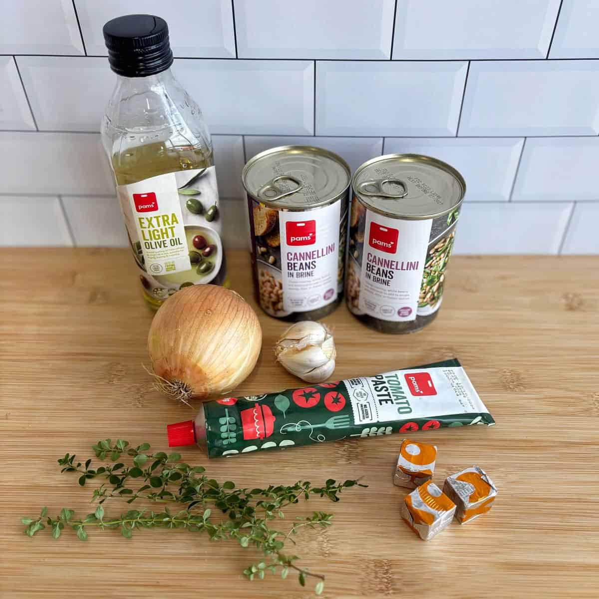 Homemade Baked Beans A bottle of olive oil, two cans of cannellini beans, an onion, a garlic bulb, a tube of tomato paste, thyme sprigs, and four vegetable stock cubes on a wooden surface.
