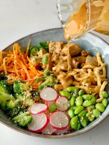 A bowl with broccoli, radish slices, edamame, shredded carrots, green onions, crispy noodles, and chicken, topped with sesame seeds and a creamy sauce being poured over.