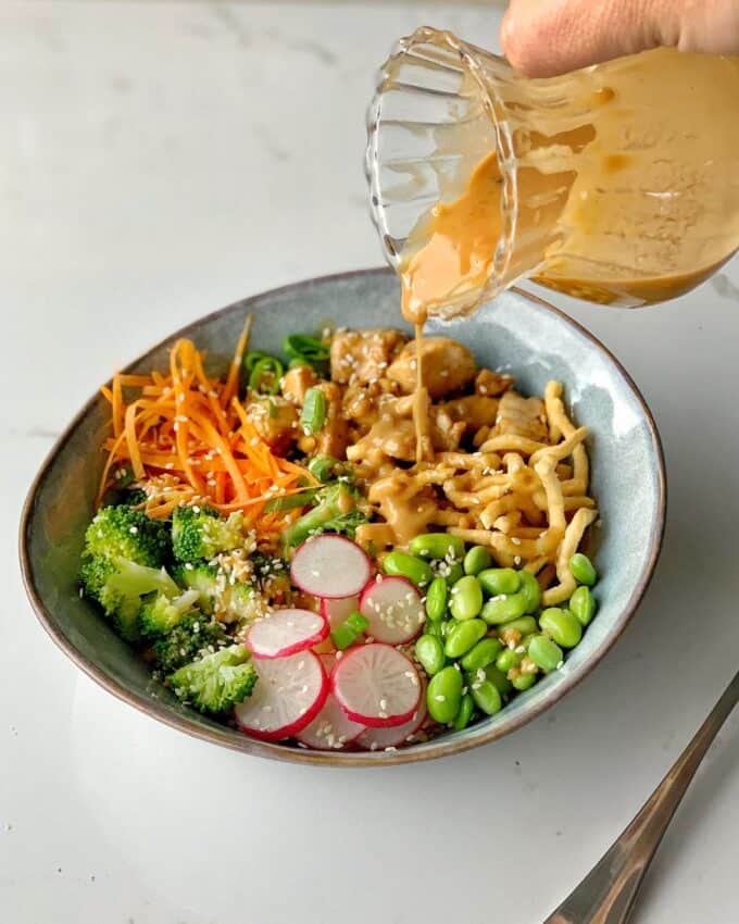 A hand pours a creamy dressing over a bowl filled with noodles, broccoli, sliced radishes, shredded carrots, edamame, and sesame seeds.