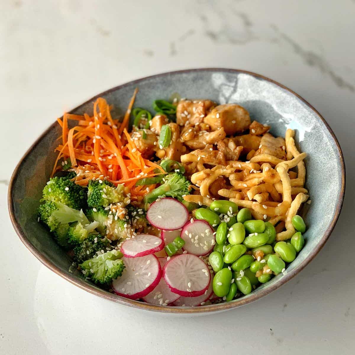 A bowl with cooked chicken, shredded carrots, broccoli, sliced radishes, snap peas, edamame, and crispy noodles, topped with sesame seeds.
