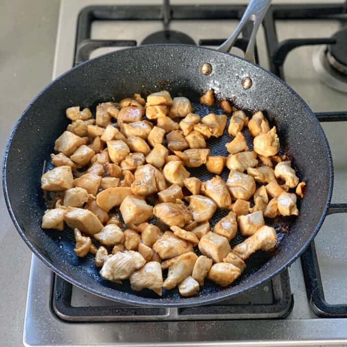 A frying pan on a stovetop contains cooked, browned chicken pieces.