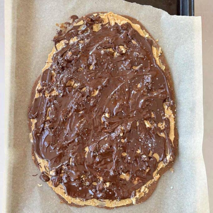 Date and Chocolate Bark A large, oval-shaped cookie topped with a layer of melted chocolate and sprinkled with salt, placed on parchment paper.