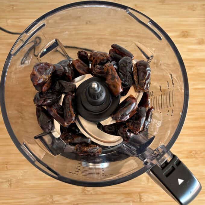 Date and Chocolate Bark A food processor containing several pitted dates on a wooden surface, viewed from above.