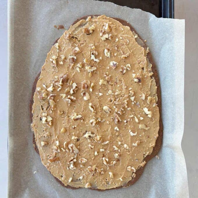 Date and Chocolate Bark Oval-shaped baked good on parchment paper, topped with a light brown spread and sprinkled with chopped nuts.