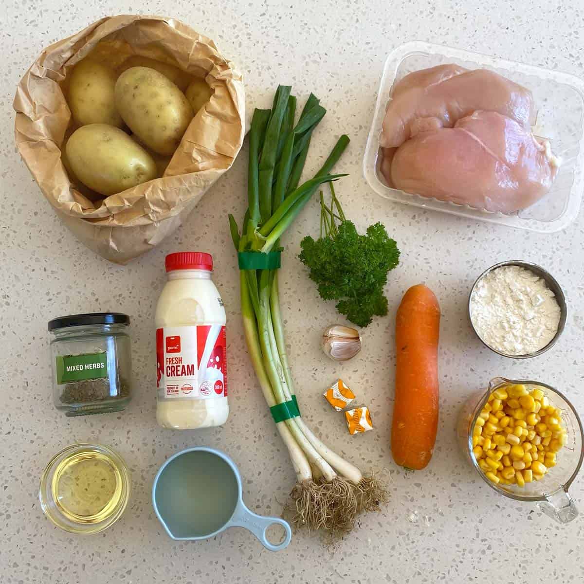 Dixie Chicken Ingredients on a counter: potatoes, chicken breasts, green onions, parsley, carrot, garlic, corn, cream, flour, mixed herbs, oil, milk, and stock cubes.