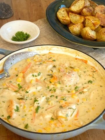 A pot of creamy chicken and vegetable stew garnished with herbs sits beside a plate of roasted potatoes on a wooden table.