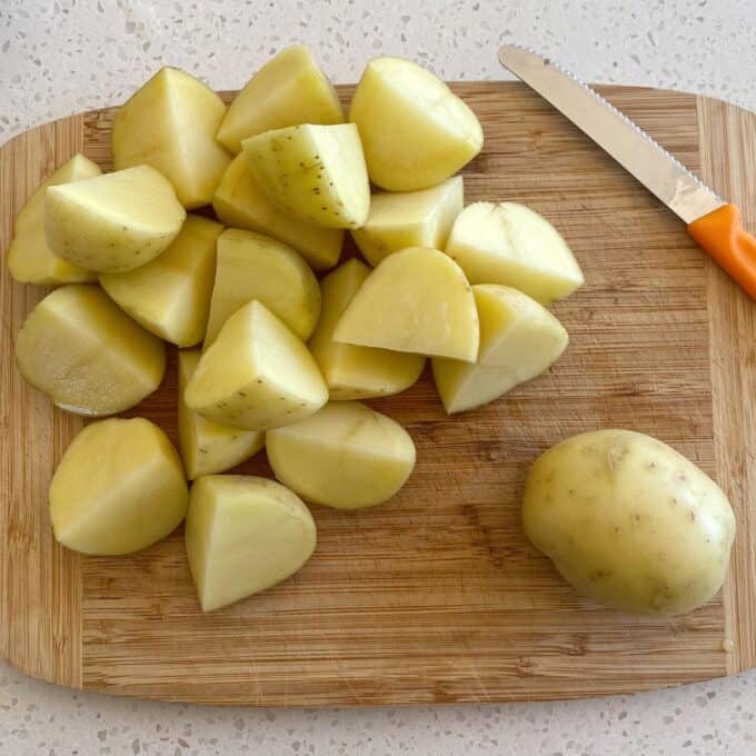 Dixie Chicken A wooden cutting board with chopped and whole potatoes, and a serrated knife with an orange handle on a light countertop.