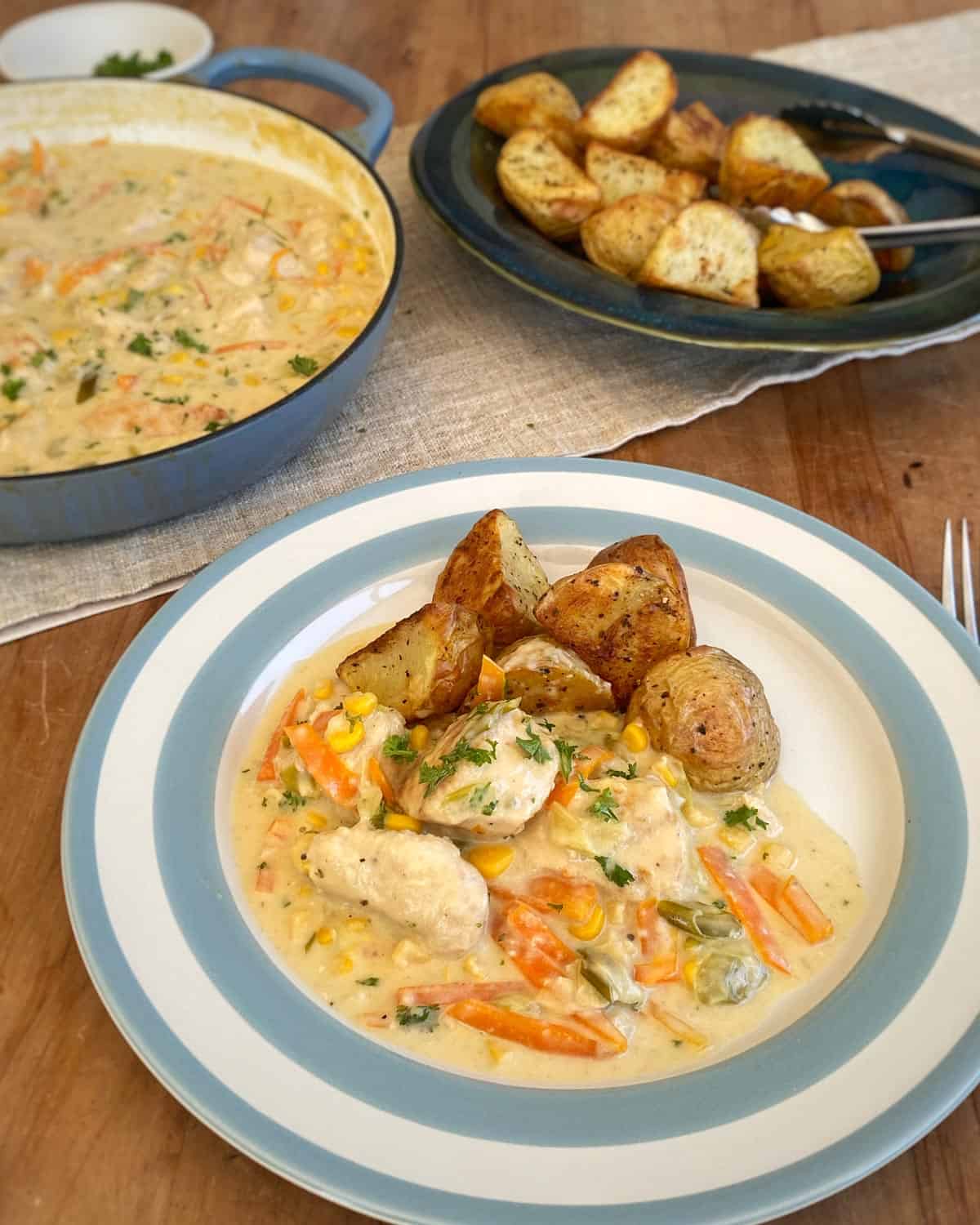 Dixie Chicken A plate of creamy chicken stew with vegetables and roasted potatoes, with a larger pot of stew and a dish of more roasted potatoes in the background.