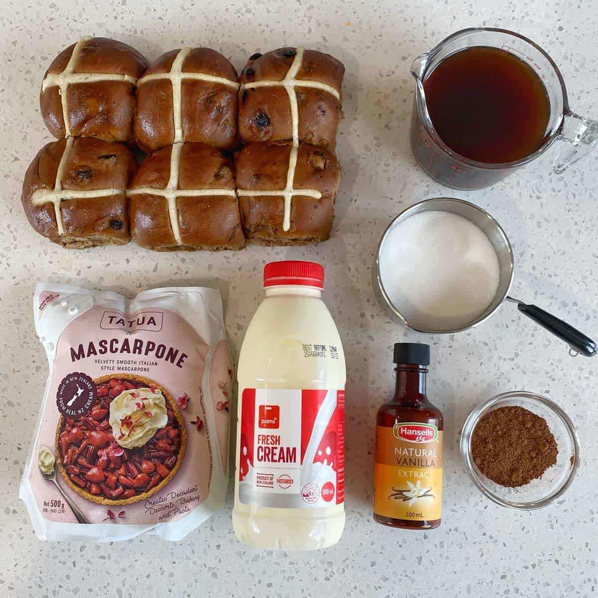 Ingredients neatly arranged on a countertop: hot cross buns, mascarpone, fresh cream, vanilla extract, cocoa powder, sugar, and a measuring jug of liquid.