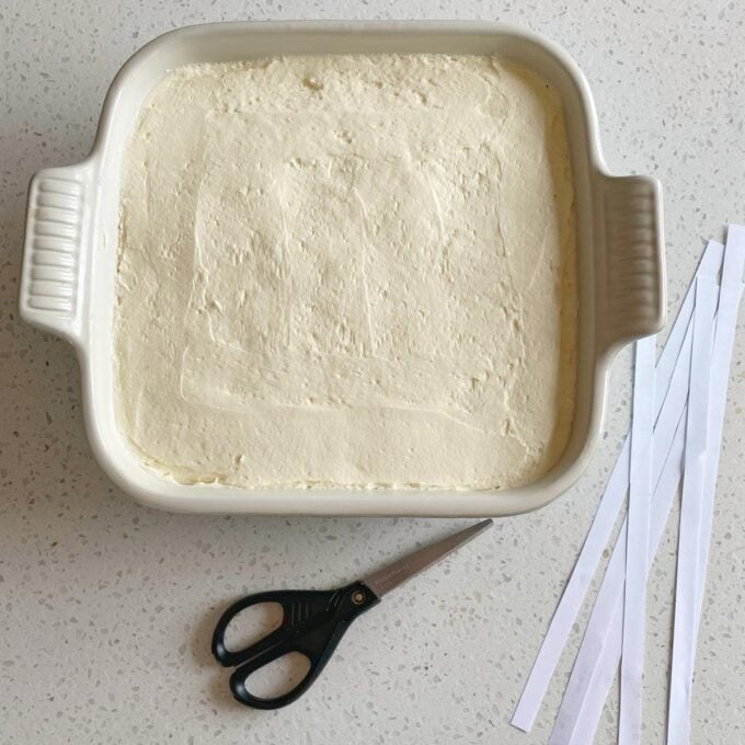 A square white dish filled with a smooth, light-colored mixture is on a countertop beside a pair of scissors and several strips of white paper.