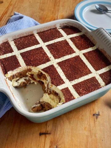 A square dish of tiramisu with a grid pattern on top, a serving missing from one corner, on a wooden table with plates, spoons, and a blue cloth nearby.