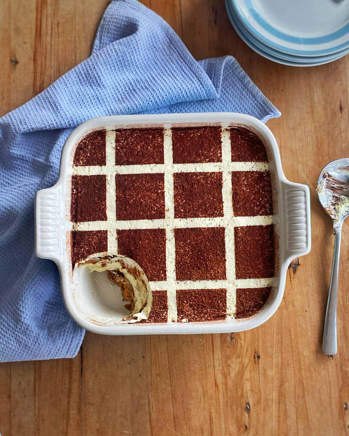 A square tiramisu in a white baking dish with a grid pattern on top, one corner scooped out, resting on a blue cloth next to a spoon and stacked plates.