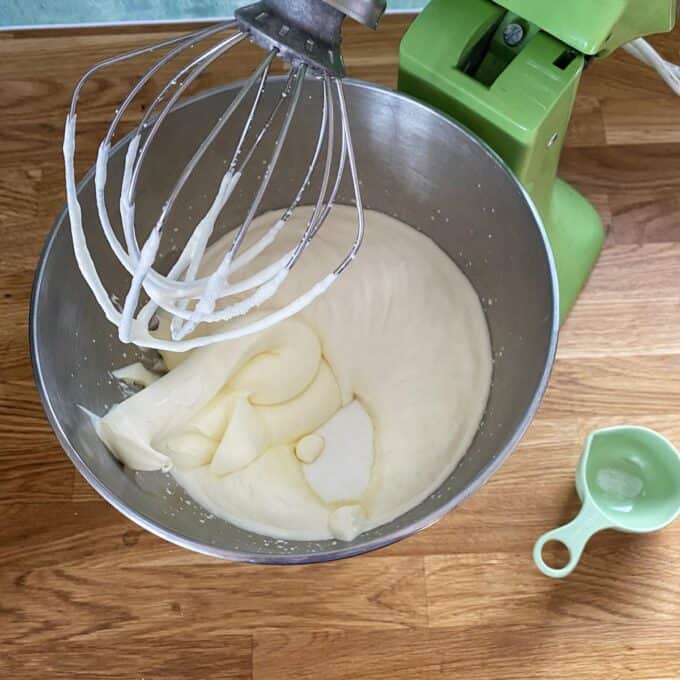A mixing bowl with thick, whipped cream and a whisk attachment, next to a green stand mixer and a small measuring cup on a wooden surface.