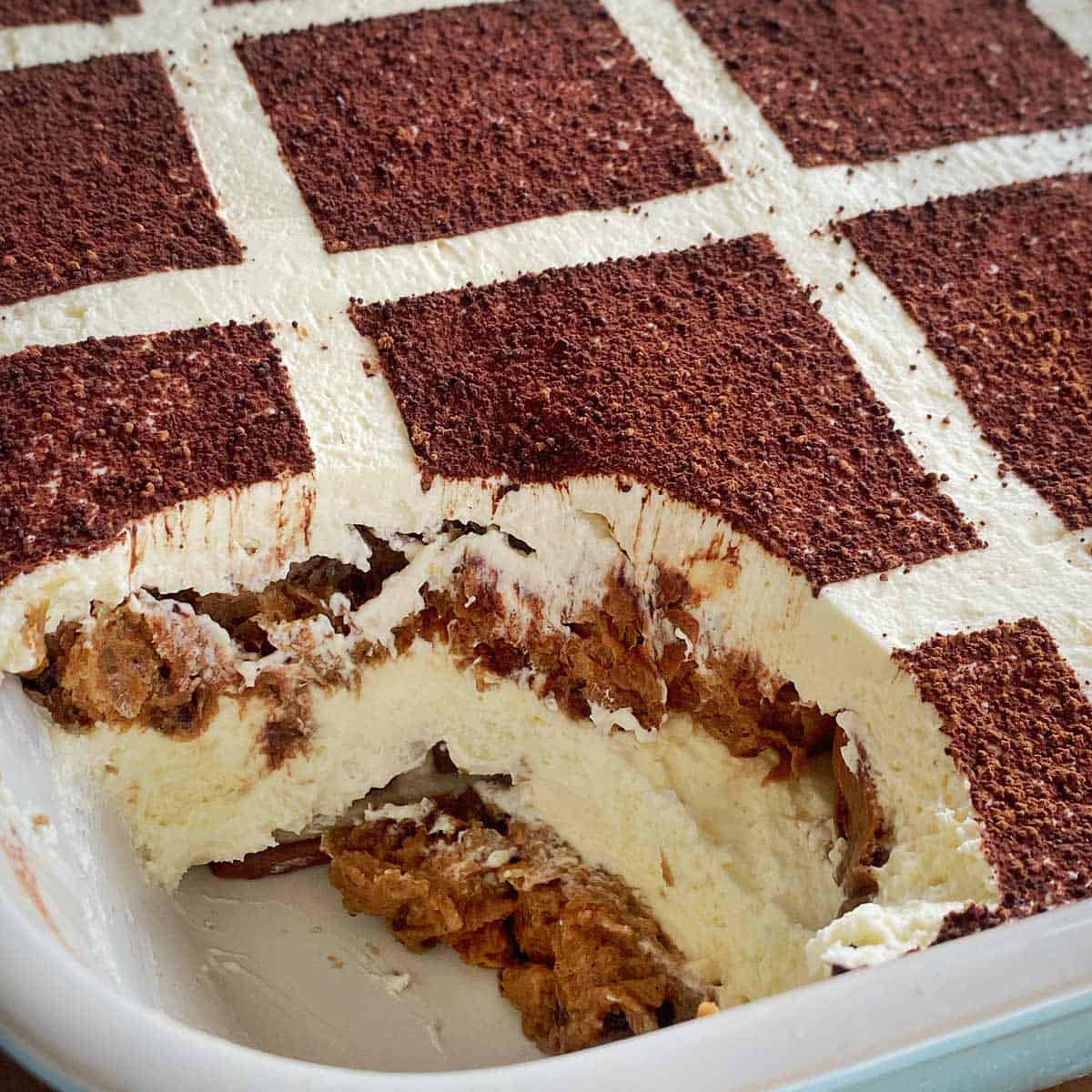 A rectangular tiramisu in a dish with a checkered cocoa powder pattern on top and a portion scooped out, revealing creamy layers inside.