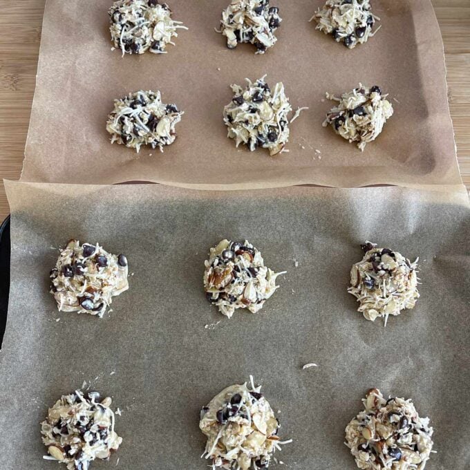 Magic Cookies Two baking trays lined with parchment paper, each holding unbaked cookie dough mounds with visible chocolate chips and shredded coconut.