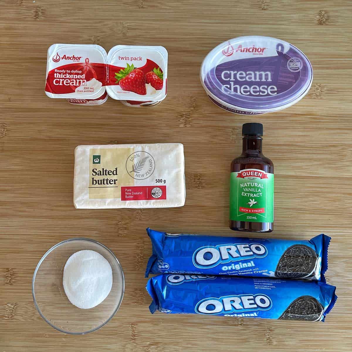 Various ingredients for a dessert are arranged on a wooden surface, including cream, cream cheese, butter, vanilla extract, sugar, and two packs of Oreo cookies.