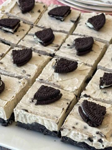 A tray of Oreo cheesecake bars, each square topped with a piece of Oreo cookie, arranged in a grid on a white plate.