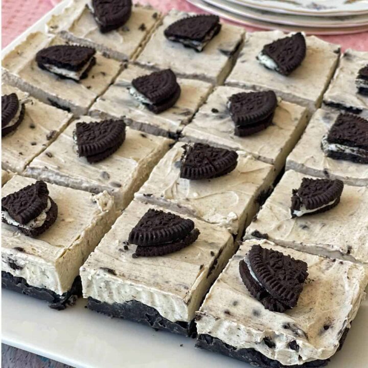A tray of Oreo cheesecake bars, each square topped with a piece of Oreo cookie, arranged in a grid on a white plate.
