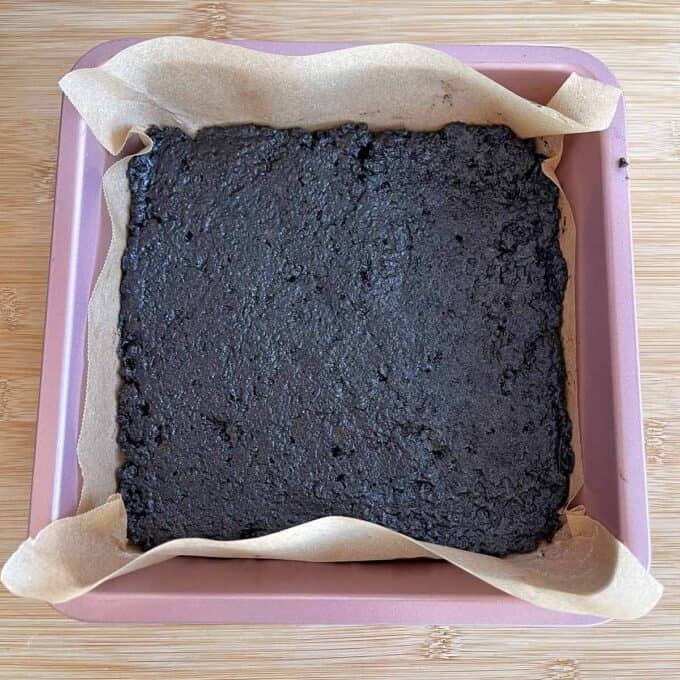 A square baking pan lined with parchment paper holds a baked, dark brown dessert, possibly brownies, resting on a wooden surface.