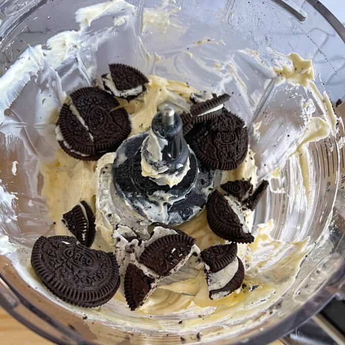 A food processor bowl contains cream cheese and several broken Oreo cookies, ready to be blended together.