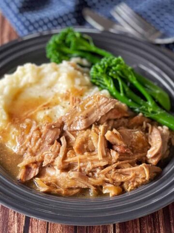 A plate of shredded roast meat with gravy, mashed potatoes, and steamed broccolini on a dark plate with a fork and knife in the background.
