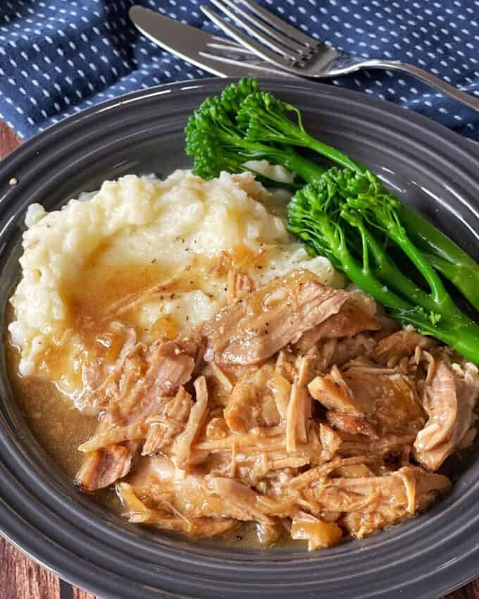 Slow Cooker Honey Pork A plate with mashed potatoes and gravy, shredded meat in sauce, and steamed broccolini, with a fork and knife on a blue patterned napkin in the background.