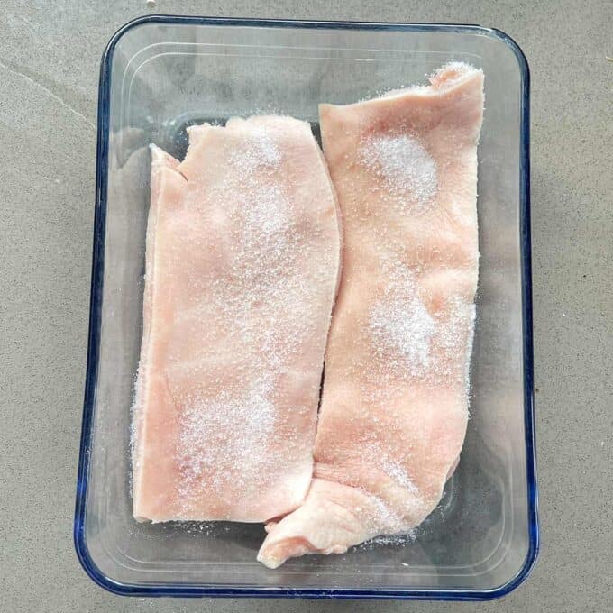 Slow Cooker Honey Pork Two pieces of raw pork skin sprinkled with salt are placed side by side in a clear rectangular glass container.