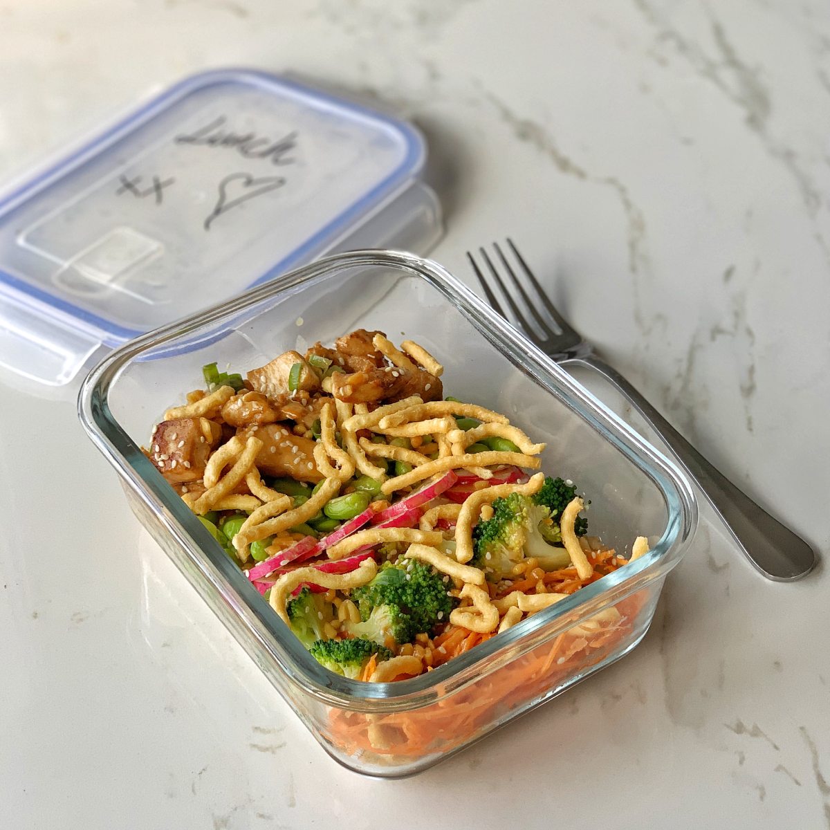 A glass container with stir-fried noodles, vegetables, and tofu is placed on a marble surface beside a fork and a labeled plastic lid.