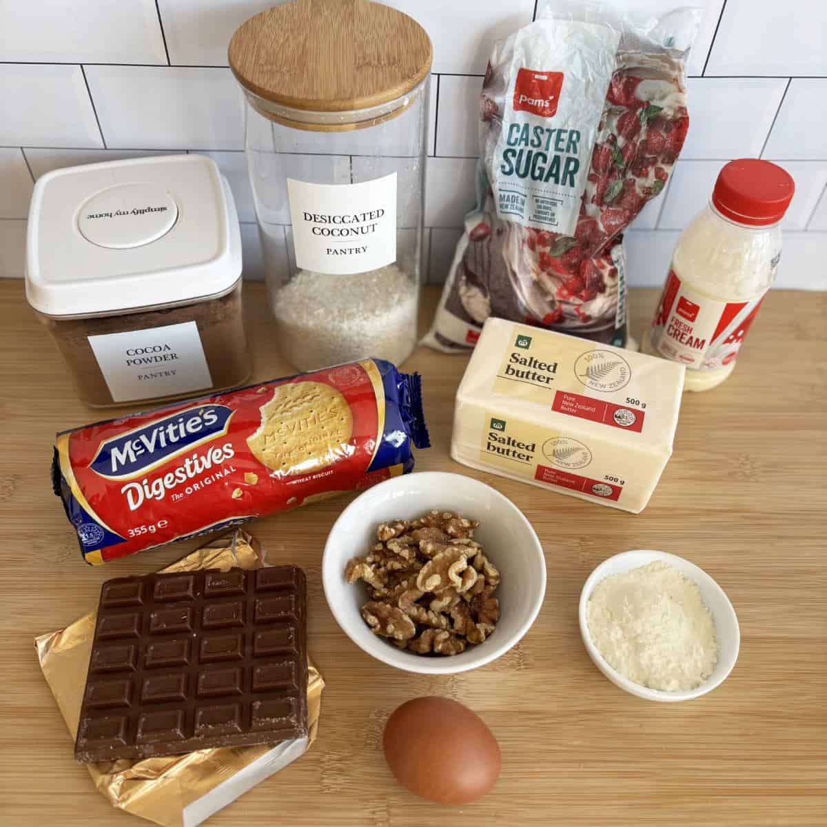 Nanaimo Slice A selection of baking ingredients on a wooden surface, ready to create a delicious Nanaimo slice—featuring chocolate, biscuits, walnuts, cocoa powder, desiccated coconut, sugar, cream, butter, and an egg.