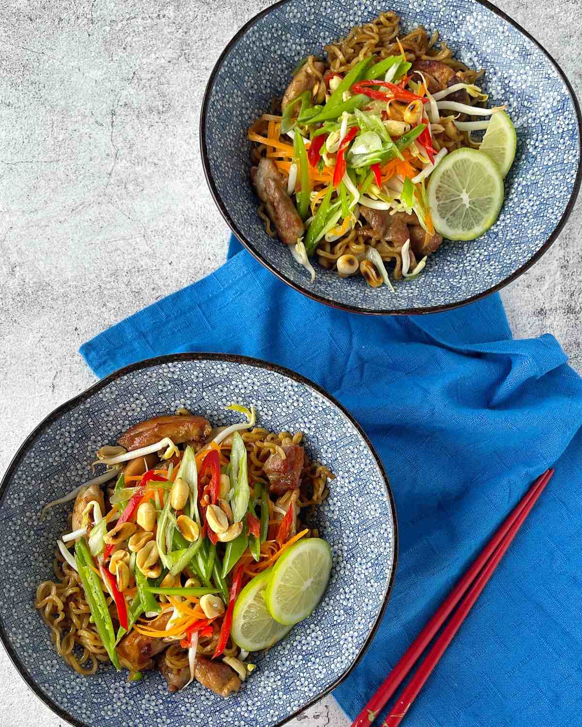 Two bowls of noodle stir-fry topped with sliced vegetables, peanuts, and lime slices on a blue cloth, with red chopsticks beside them.