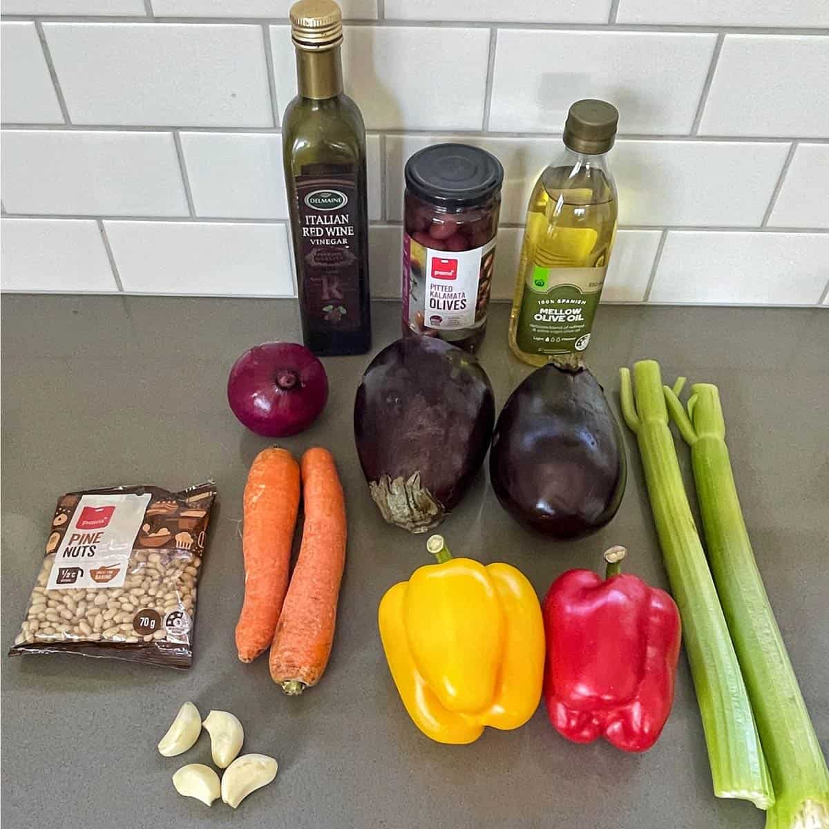 Caponata Assorted vegetables, pine nuts, two bottles of oil, a jar of olives, and garlic cloves arranged on a kitchen counter with a white tile backsplash.
