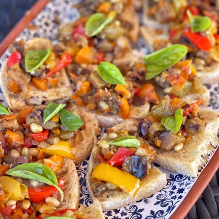 A platter of toasted bread slices topped with mixed roasted vegetables, pine nuts, and fresh basil leaves, arranged on a patterned serving tray.