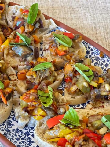A decorative platter with slices of bread topped with a vegetable mixture, pine nuts, and fresh basil leaves, arranged on a patterned plate.