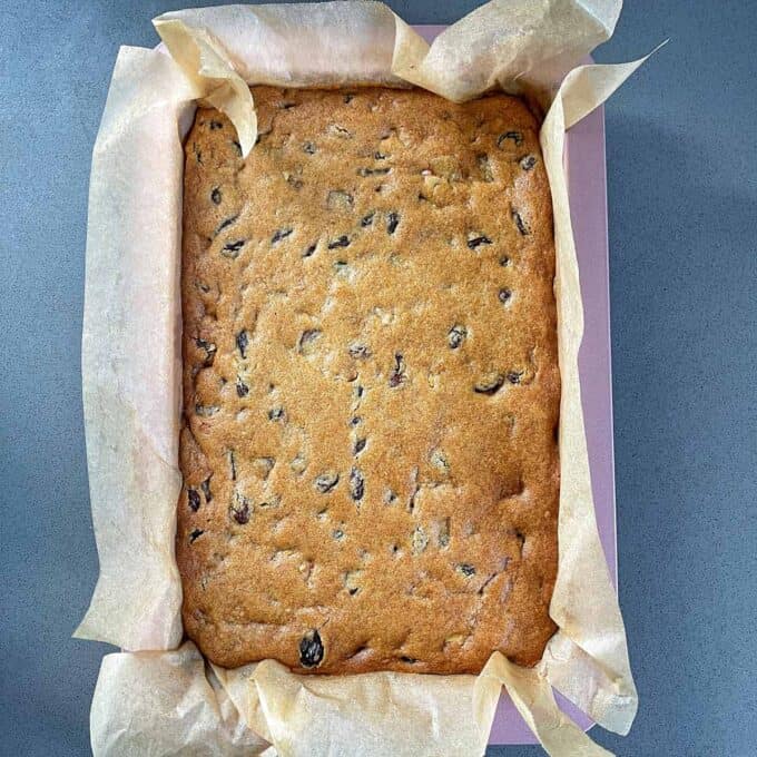 Golden Syrup Sultana Chews A rectangular fruit cake baked in a parchment paper-lined pan, with a golden-brown top and visible pieces of dried fruit.
