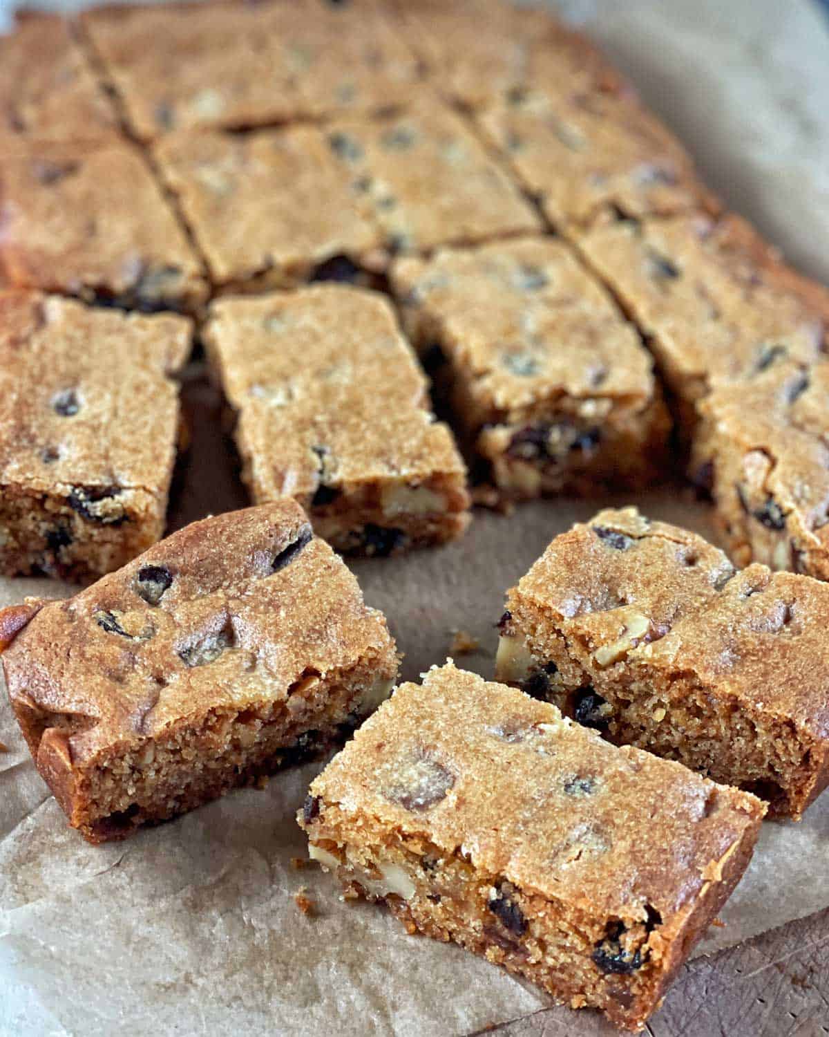 Golden Syrup Sultana Chews A batch of rectangular, golden-brown cake bars with nuts and chocolate chips, sliced and arranged on parchment paper.