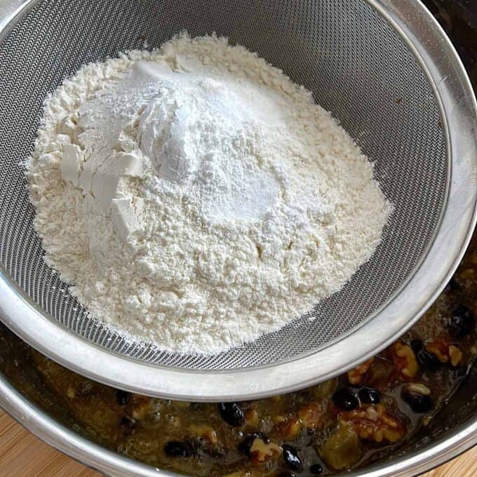 Golden Syrup Sultana Chews Flour and baking powder in a metal sifter positioned above a bowl containing a mixture of nuts, dried fruit, and liquid ingredients.