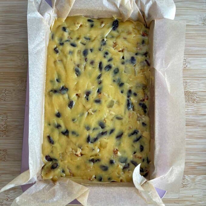 Golden Syrup Sultana Chews Unbaked fruitcake batter with visible raisins and nuts spread in a parchment-lined rectangular baking tin on a wooden surface.