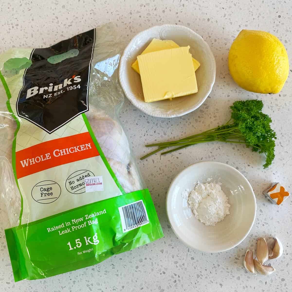 Lemon Garlic Butter Roast Chicken Whole raw chicken in packaging, a bowl of butter slices, a lemon, parsley, a bowl of salt, chicken stock cube, and garlic cloves on a countertop.