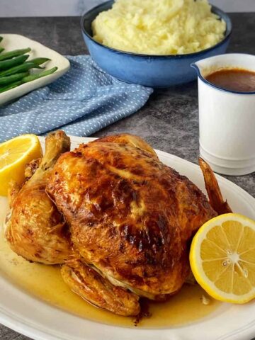Roast chicken garnished with lemon halves on a platter, served with a side of green beans, mashed potatoes in a bowl, and a jug of gravy.