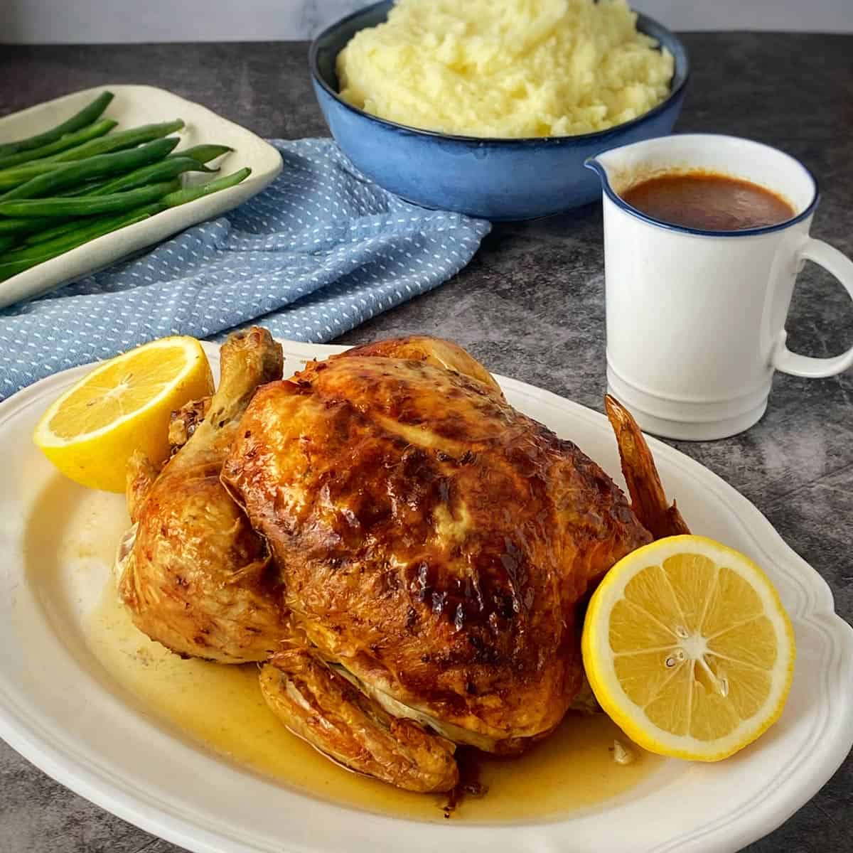 Lemon Garlic Butter Roast Chicken Roast chicken garnished with lemon halves on a platter, served with a side of green beans, mashed potatoes in a bowl, and a jug of gravy.