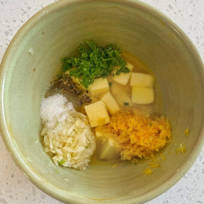 Lemon Garlic Butter Roast Chicken A green bowl containing chopped garlic, lemon zest, butter cubes, chopped parsley, salt, pepper, and olive oil on a light countertop.